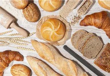 A variety of fresh bread, including croissants and rolls, is laid out on a wooden tray. Also arranged are wheat grains and a rolling pin.