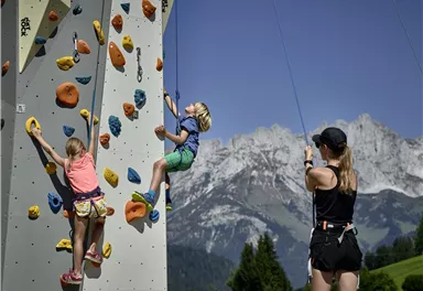 Ein Kletterturm in den Bergen, wo Kinder klettern. Im Hintergrund sind beeindruckende Berglandschaften zu sehen.