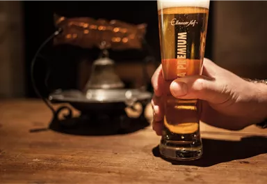 A glass of beer is held by a hand. In the background, an antique lamp is visible.