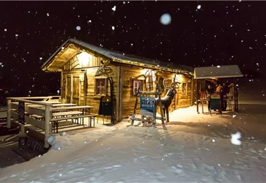 A cozy mountain cabin in the snow at night, with festive lighting. In the foreground, tables and a figure of a deer are visible.