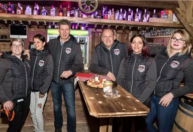A group of six people is standing in a cozy bar with wood decor. They are wearing dark gray vests and smiling warmly at the camera.