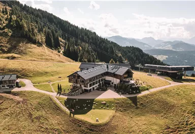 Ein malerisches Bergrestaurant umgeben von grünen Wiesen und bewaldeten Hügeln. Im Hintergrund sind die majestätischen Berge zu sehen.