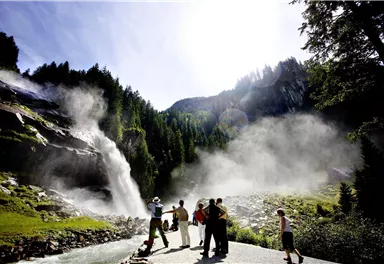A group of people is standing near an impressive waterfall surrounded by green trees. The sky is clear and the sun is shining brightly.