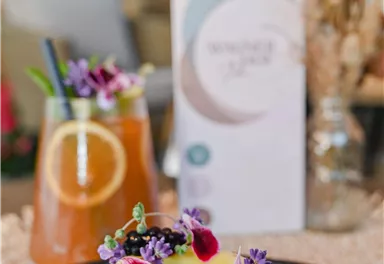 A delicious cake with a dish made of fresh herbs and edible flowers. In the background, two drinks and a menu card are visible.