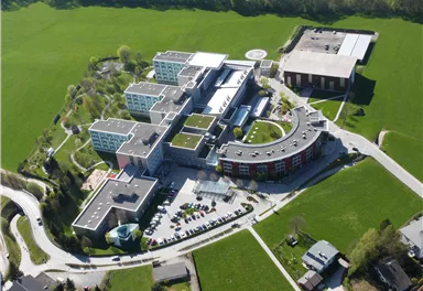 Ein modernes Gebäudeensemble in einer grünen Landschaft. Die Struktur umfasst mehrere separate Gebäude und ist von Rasenflächen umgeben.