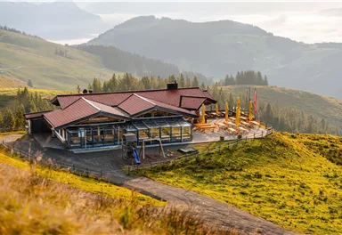 Ein modernes Gebäude in den Bergen mit einer Terrasse und weiten Grünflächen. Im Hintergrund sind sanfte Hügel und ein klarer Himmel zu sehen.