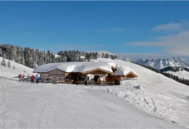 Eine gemütliche Berghütte liegt in einer verschneiten Winterlandschaft. Im Hintergrund sind schneebedeckte Berge und ein klarer blauer Himmel zu sehen.