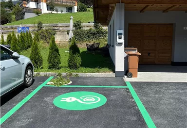 Ein Parkplatz mit einem grünen Elektrolade-Symbol. Im Hintergrund sind Häuser und Bäume zu sehen.