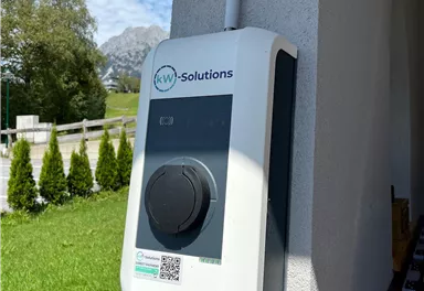 Eine Elektro-Ladestation an einer Wand, umgeben von Grünflächen und Bäumen. Der Himmel ist blau mit einigen Wolken.