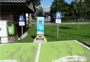Ein Parkplatz für Elektrofahrzeuge mit einer Ladestation und sichtbaren Markierungen. Im Hintergrund befinden sich weitere Parkplätze und ein grüner Bereich.