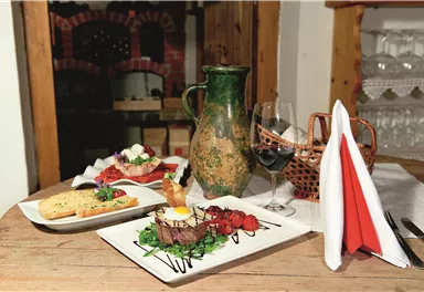 Ein schön gedeckter Tisch mit verschiedenen Gerichten, darunter ein Steak auf grünen Blättern, ein Salat und ein Stück Brot. Dazu sind ein Weinglas und eine dekorative Karaffe zu sehen.