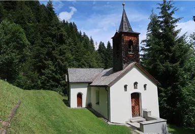A small church surrounded by dense trees. The sky is blue with some clouds.