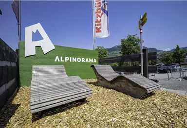 A welcome sign with the inscription "Alpinorama" and two modern wooden loungers. The surroundings are decorated with bark mulch and feature a clear blue sky.