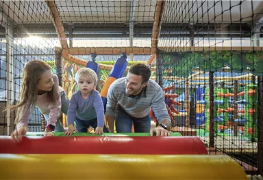 Ein Kinderspielplatz im Innenbereich mit bunten Spielgeräten. Ein Vater spielt mit seinen zwei Kindern, die auf einem farbigen Hindernisparcours klettern.