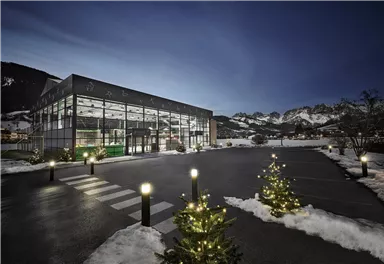 Ein modernes Gebäude in einer verschneiten Landschaft. Umgeben von beleuchteten Weihnachtsbäumen und Bergen im Hintergrund.