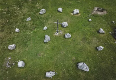 A green meadow with several large stones and a cross in the middle. In the background, some more stones and a small seating area are visible.