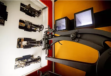 Ein historischer Drucker steht im Vordergrund, umgeben von alten Druckmaschinen an der Wand. Die Maschinen zeigen verschiedene Drucktechniken und erinnern an die Geschichte des Druckens.
