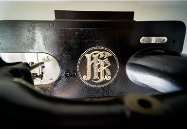 Eine klassische Nähmaschine mit einem dekorativen Logo in Schwarz und Gold. Die Maschine zeigt feine Details und handwerkliche Kunstfertigkeit.