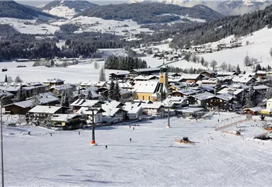 Eine malerische Winterlandschaft mit einem verschneiten Dorf. Die Umgebung ist von Bergen und Bäumen umgeben, während Skifahrer auf der Piste sind.
