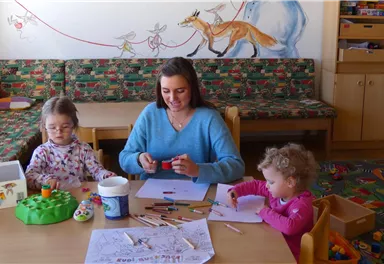 A woman is sitting at a table with two little girls, crafting. Around her are various crafting materials and a friendly, colorful room.