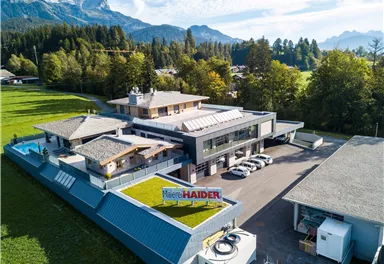 Ein modernes Gebäude in einer ländlichen Umgebung, umgeben von Bäumen und Bergen. Es gibt Parkplätze und eine große Wiese im Vordergrund.