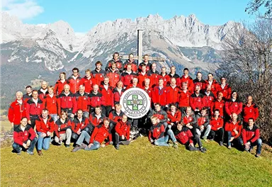 Eine große Gruppe von Menschen in roten Jacken steht auf einer Wiese vor majestätischen Bergen. Sie posieren um ein gemeinsames Schild in der Mitte des Bildes.
