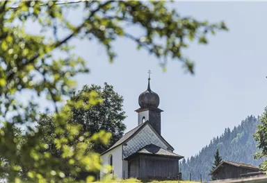 A picturesque church stands among trees with a mountain in the background. The sky is clear and sunny.