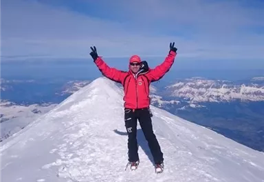 Eine Person steht auf einem schneebedeckten Gipfel und jubelt. Im Hintergrund sind die Berge und ein klarer Himmel sichtbar.