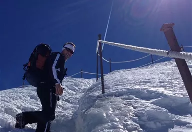 Ein Bergsteiger klettert einen schneebedeckten Hang hinauf. Über ihm strahlt die Sonne in einem klaren blauen Himmel.