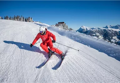 A skier in red clothing is skiing over the snow in the mountains. The clear blue sky and the snow-capped peaks in the background create a beautiful winter landscape.