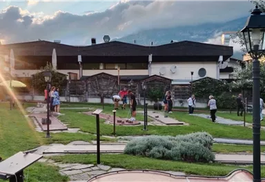 Eine Mini-Golfanlage mit mehreren Spielern im Freien. Im Hintergrund sind Bäume und ein Gebäude zu sehen.