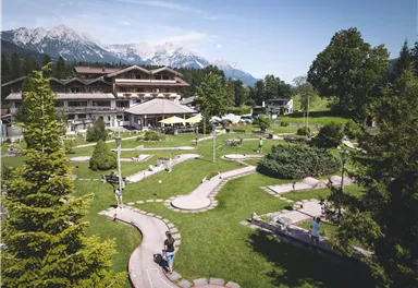Eine großzügige Gartenlandschaft mit einem Mini-Golfplatz und verschiedenen Bäumen. Im Hintergrund sind Berge und ein stilvolles Gebäude zu sehen.