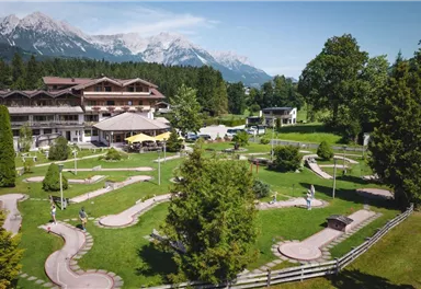 Eine schöne Mini-Golfanlage mit gepflegten Bahnen und viel Grün. Im Hintergrund sind Berge und ein Hotel zu sehen.