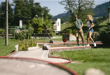 Ein Mini-Golfplatz mit grüner Rasenfläche und roten Markierungen. Zwei Personen gehen entspannt entlang der Spielbahnen.
