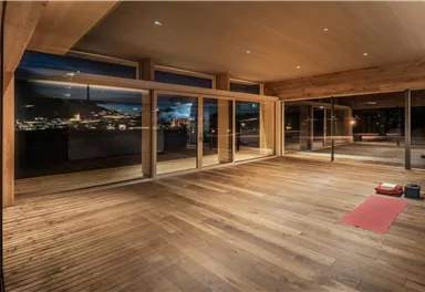A modern, light-filled room with wooden flooring and large windows. In the foreground lies a red yoga mat that creates a relaxing atmosphere.