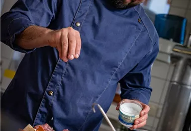 Ein Koch in einer modernen Küche würzt frisches Gemüse und Fleisch. Er trägt eine blaue Kochjacke und lächelt beim Zubereiten des Essens.