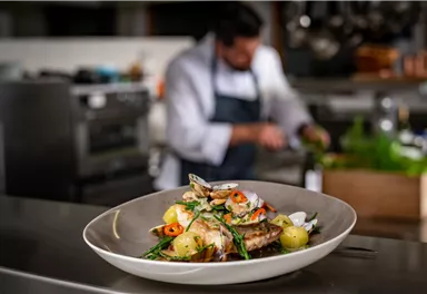Ein köstliches Gericht mit Fisch, Kartoffeln und frischen Kräutern steht auf einem Tisch. Im Hintergrund arbeitet ein Koch in der Küche.