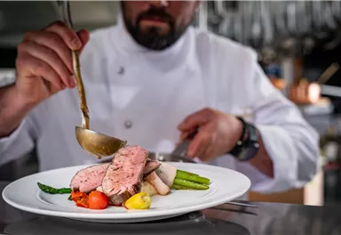 Ein Koch richtet ein schönes Gericht mit rosa gebratenem Fleisch und frischem Gemüse an. Der Fokus liegt auf der Präsentation des Essens auf einem weißen Teller.