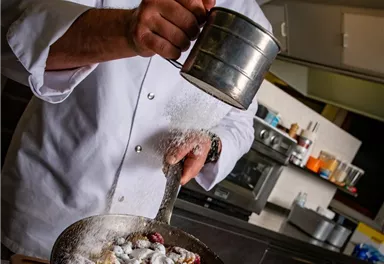 Ein Küchenchef streut Puderzucker über eine frische Obstschüssel. Im Hintergrund sind Kochutensilien und eine moderne Küche zu sehen.