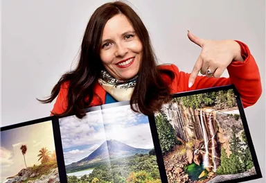 A smiling woman holds a photo book with pictures of nature and waterfalls. She points to the impressive landscapes in the book.