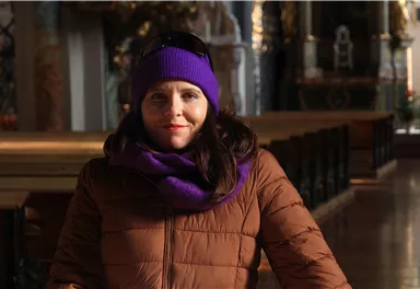 A woman with long hair is wearing a purple hat and a brown coat. She is standing in a decorative interior with wooden details.