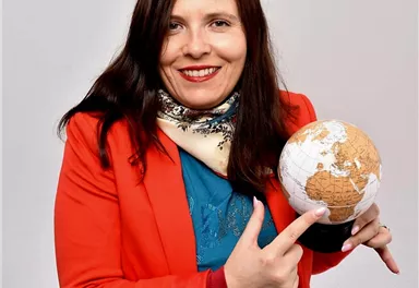 A smiling woman in a red blazer holds a globe in her hand. She points at the globe and is wearing a scarf around her neck.