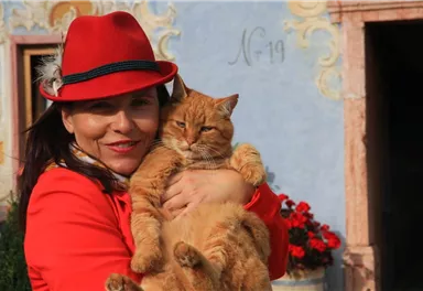 A woman with a red hat holds an orange cat in her arms. In the background, blooming plants and a colorful wall are visible.