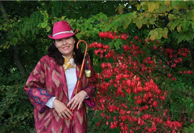 A smiling woman in a colorful outfit stands next to a blooming shrub with red fruits. She is wearing a hat and holding a walking stick in her hand.