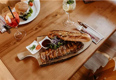 Ein köstliches Gericht aus gegrilltem Fleisch auf einem Holzbrett, serviert mit Brot und Dip. Im Hintergrund sind Cocktails und ein Salat zu sehen.
