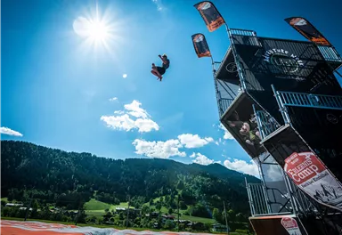 Ein Sportler springt von einer hohen Plattform in den Himmel. Im Hintergrund sind grüne Hügel und ein klarer blauer Himmel zu sehen.