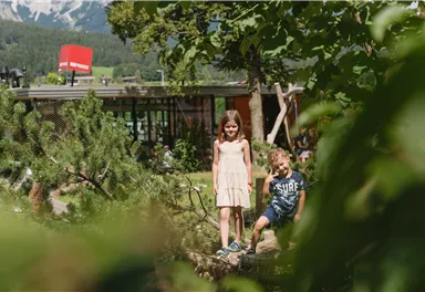 Zwei Kinder stehen in einem Garten mit Bäumen und Pflanzen. Im Hintergrund ist eine moderne Gebäudeansicht zu sehen.