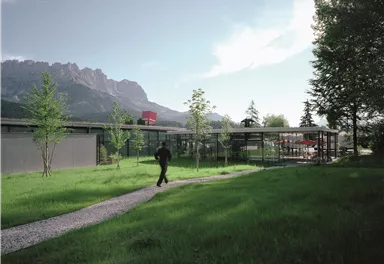 Ein modernes Gebäude in einer grünen Landschaft mit Bergen im Hintergrund. Ein Mann geht auf einem Weg entlang, der durch das Gras führt.