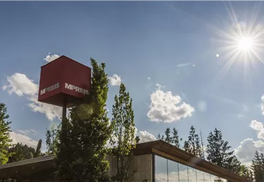 Ein modernes Gebäude mit einer großen Fensterfront und einem roten Schild. Strahlender Sonnenschein und blaue Himmel mit einigen Wolken im Hintergrund.