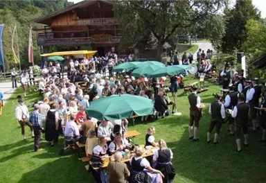 Eine große Menschenmenge versammelt sich im Freien bei einem Fest. Es gibt viele Tische, Sonnenschirme und traditionelle Kleidung.